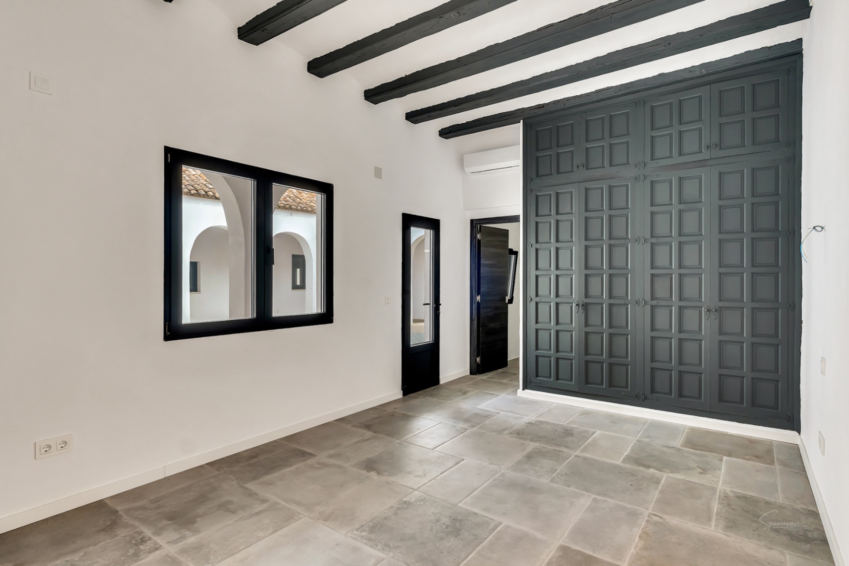 Bedroom featuring dark wooden ceiling beams, natural stone flooring, and a large panelled wardrobe. A black-framed window overlooks the traditional arched interior patio.