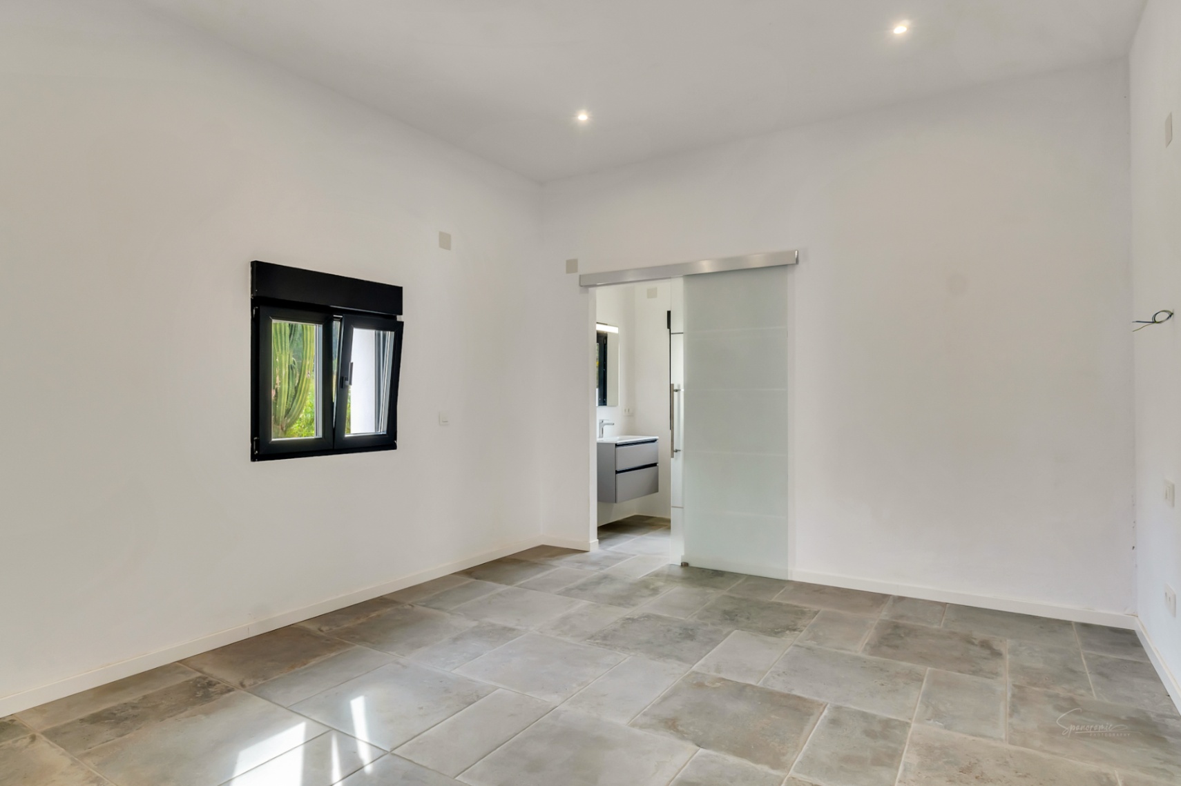 Bright bedroom featuring natural stone flooring, a black-framed window, and a frosted glass sliding door leading to a modern en-suite bathroom.