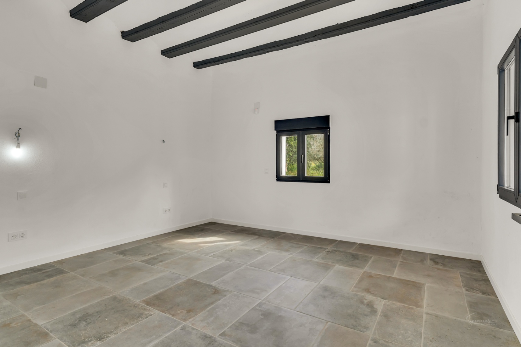 Bright, empty room featuring premium stone tile flooring, exposed dark wood ceiling beams, and black-framed windows that contrast with the clean white walls.