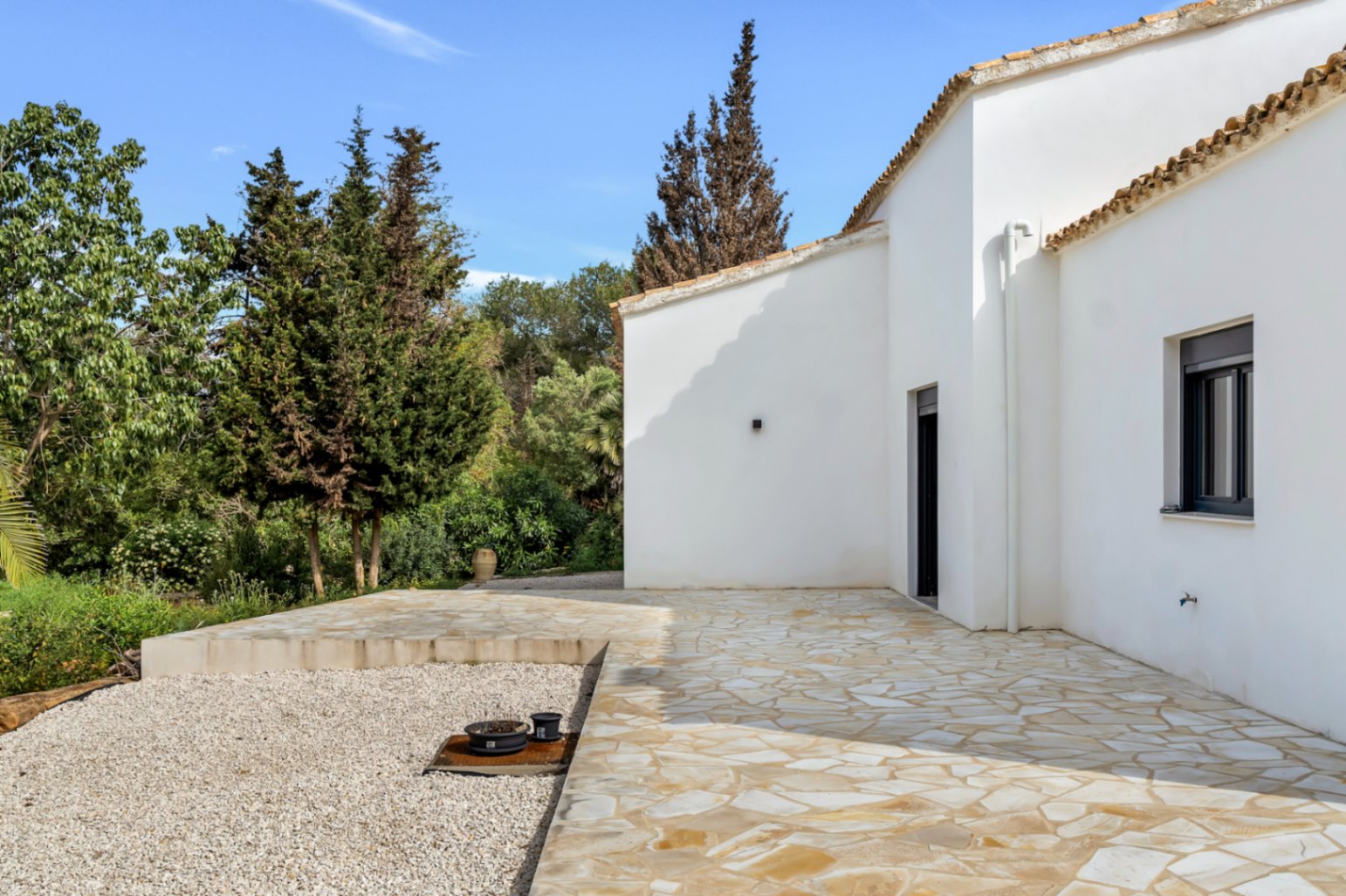 Exterior of the villa featuring white stucco walls, terracotta roofing, and a spacious natural stone terrace bordered by lush Mediterranean greenery.
