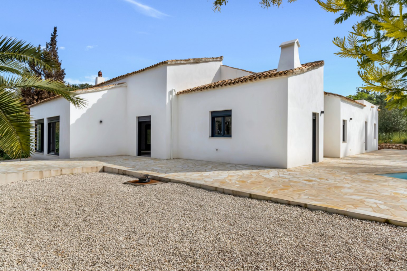 Exterior view of a Mediterranean villa featuring white stucco walls, terracotta roof tiles, and a spacious natural stone terrace overlooking a gravel area.