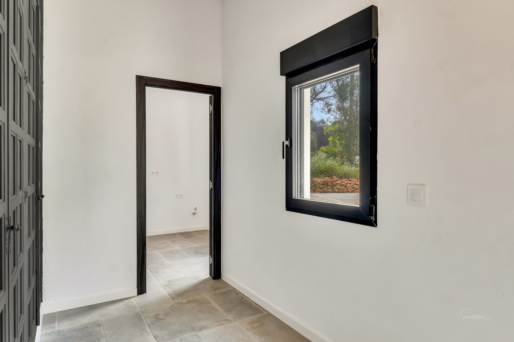Hallway featuring natural stone flooring and white walls, with dark-framed windows and doors offering a view of the surrounding greenery.