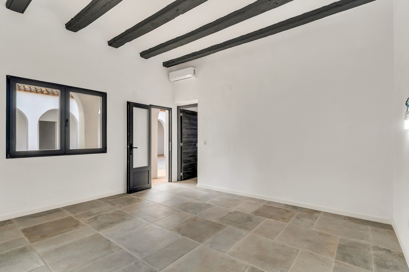 Interior room featuring exposed dark wood ceiling beams, natural stone flooring, and views of the arched central courtyard through black-framed windows.