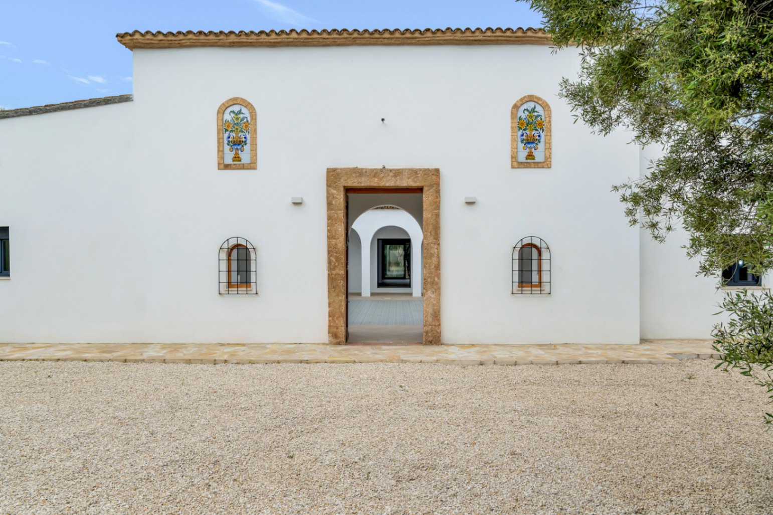Mediterranean facade featuring white stucco, Tosca stone entrance, and decorative ceramic tiles. The central doorway offers a view into the arched interior courtyard.