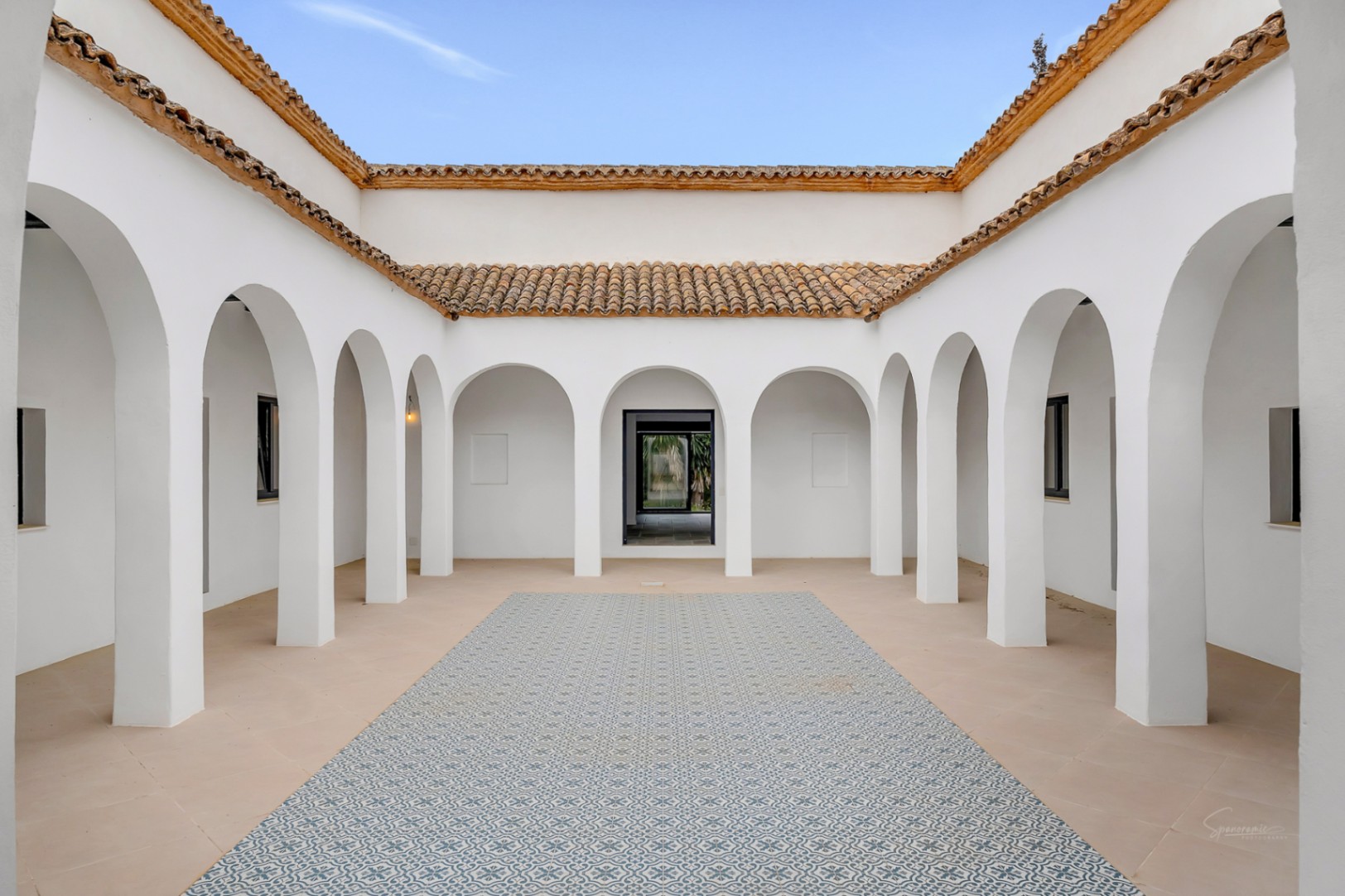 Mediterranean-style interior courtyard featuring symmetrical white arches, stone flooring with a decorative patterned tile center, and traditional terracotta roof tiles.