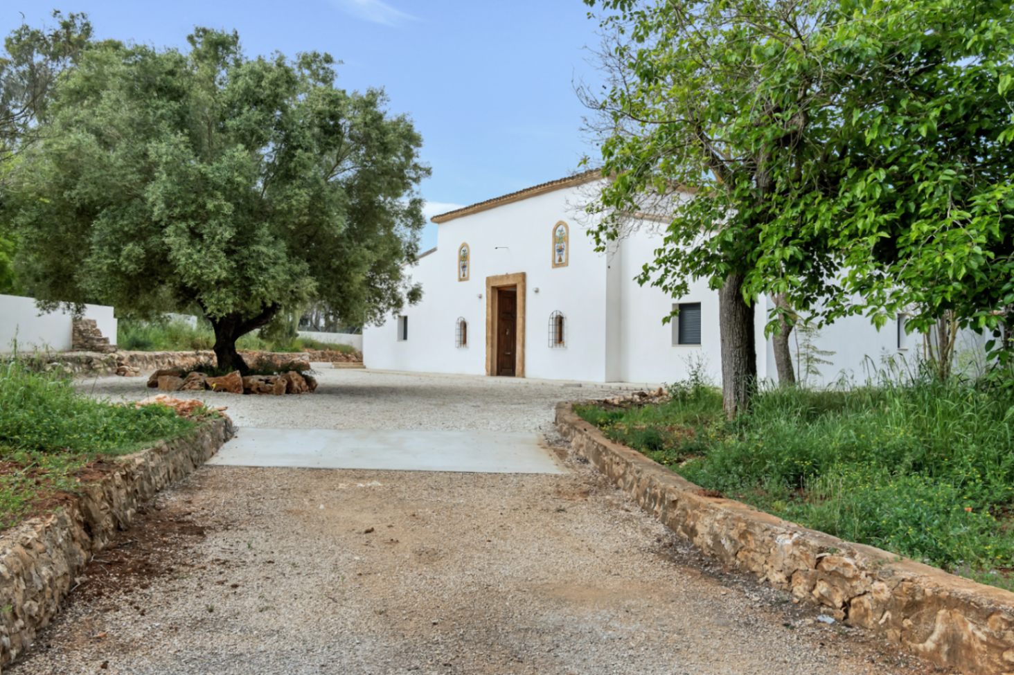 Mediterranean-style main facade featuring white stucco walls, a natural stone entrance, and a wide gravel driveway surrounded by mature olive trees and native greenery.