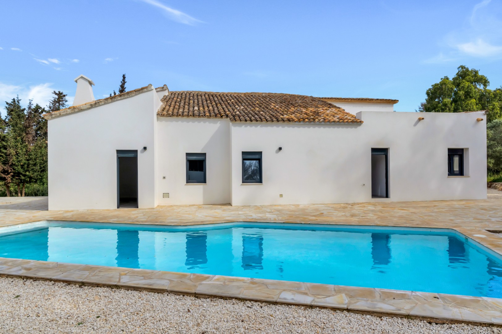 Mediterranean villa exterior featuring white stucco walls, terracotta roof tiles, and a swimming pool bordered by a natural stone terrace and gravel.