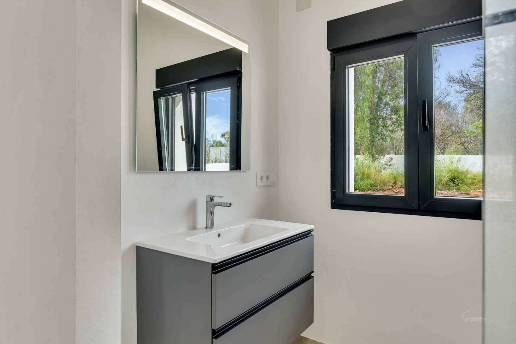Modern bathroom featuring a charcoal grey floating vanity, white ceramic sink, and LED-lit mirror. A black-framed window provides natural light and garden views.