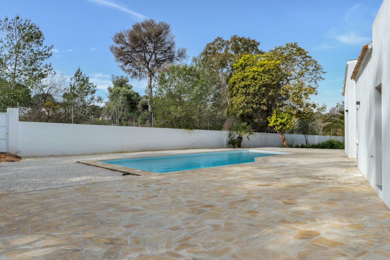 Outdoor swimming pool featuring a large natural stone terrace and white perimeter walls, set against a backdrop of mature Mediterranean trees.