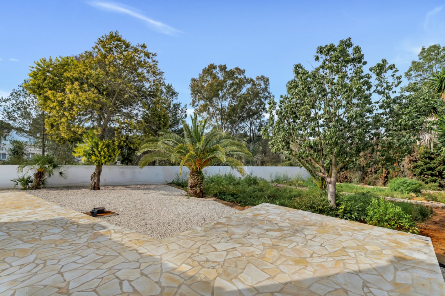 Outdoor terrace featuring irregular natural stone paving, overlooking a Mediterranean garden with a central palm tree and gravel landscaping under a clear sky.