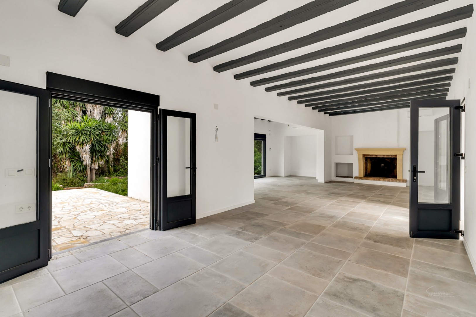 Spacious living area featuring exposed dark wooden beams, natural stone flooring, and a traditional fireplace. Large glass doors offer direct access to the stone-paved terrace.
