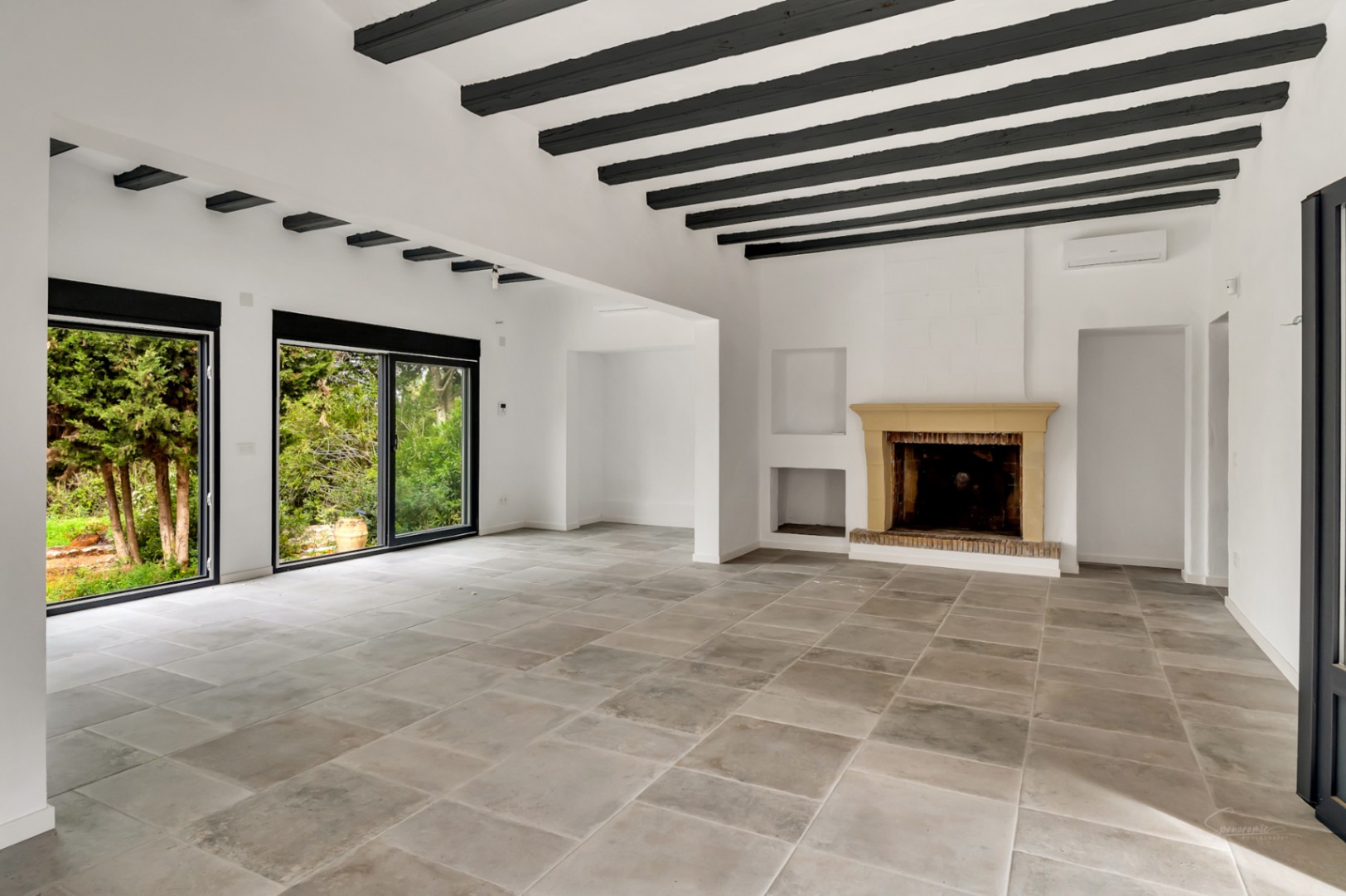 Spacious living room featuring a stone fireplace, exposed dark wood beams, and natural stone tiles. Large sliding glass doors provide seamless access to the garden.