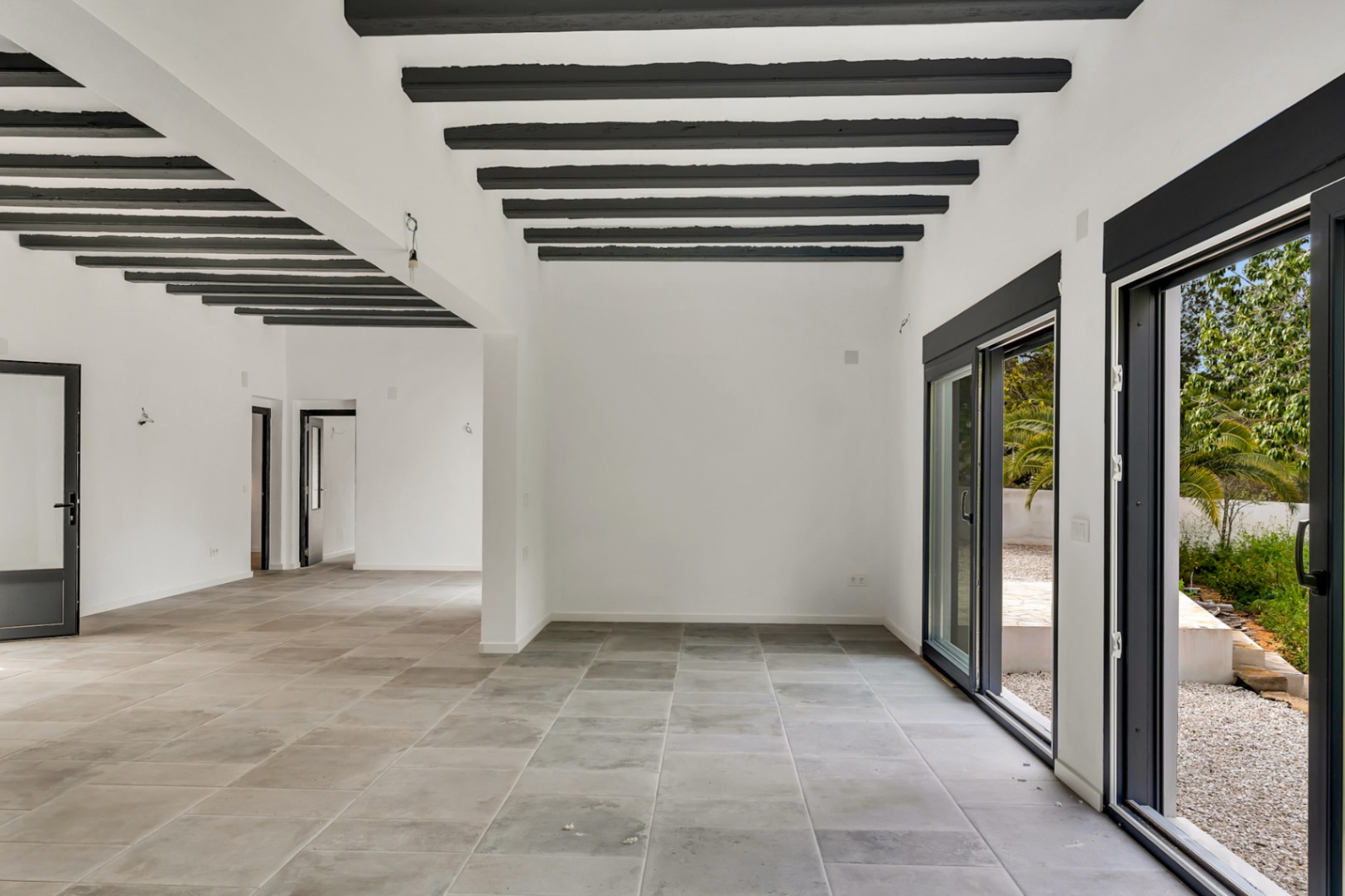 Spacious open-plan living area featuring exposed dark wooden beams, large-format stone flooring, and expansive sliding glass doors opening to the garden.
