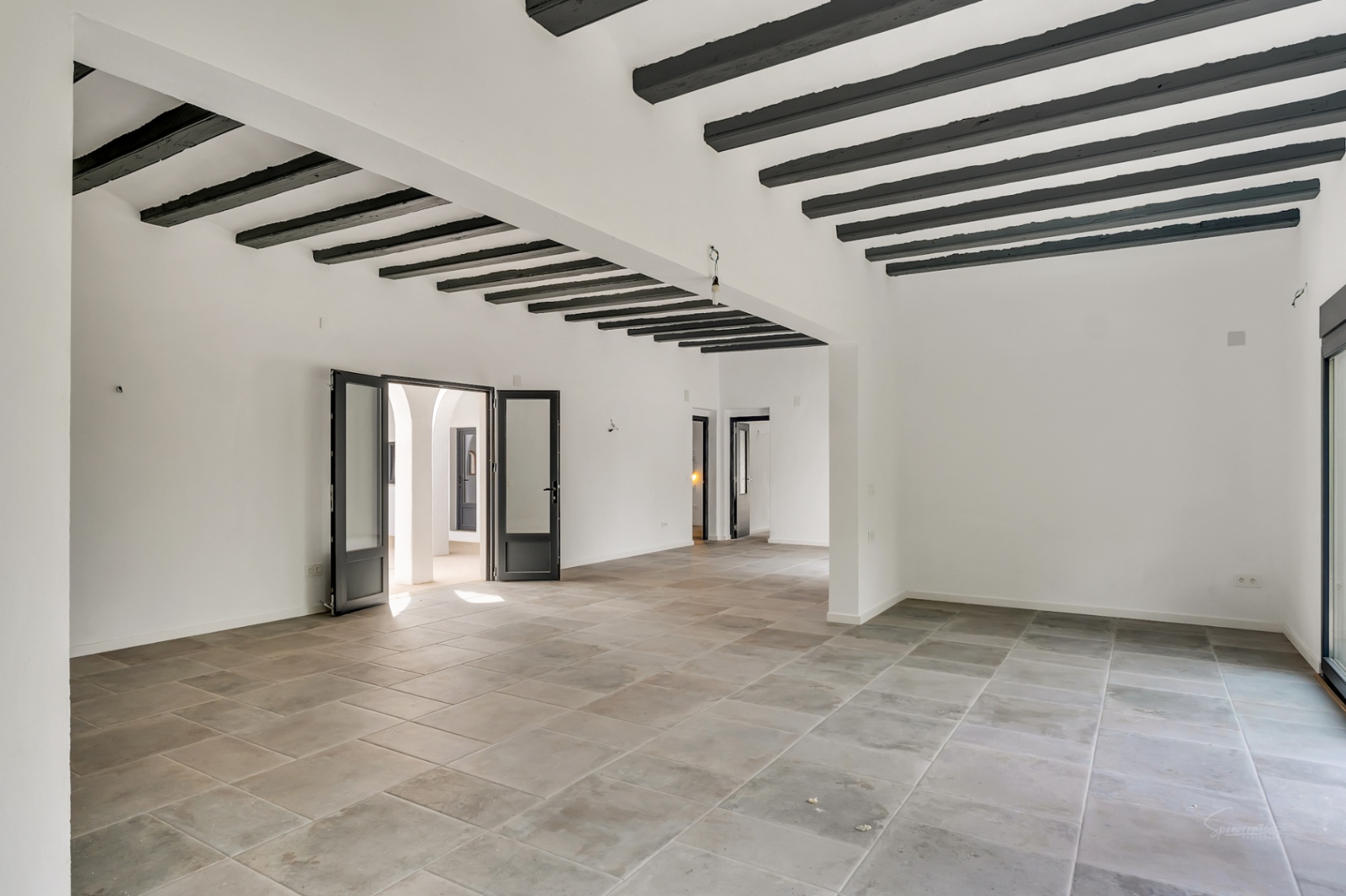 Spacious open-plan living area featuring natural stone flooring and high ceilings with exposed dark wooden beams. Large glass doors provide access to the inner courtyard.