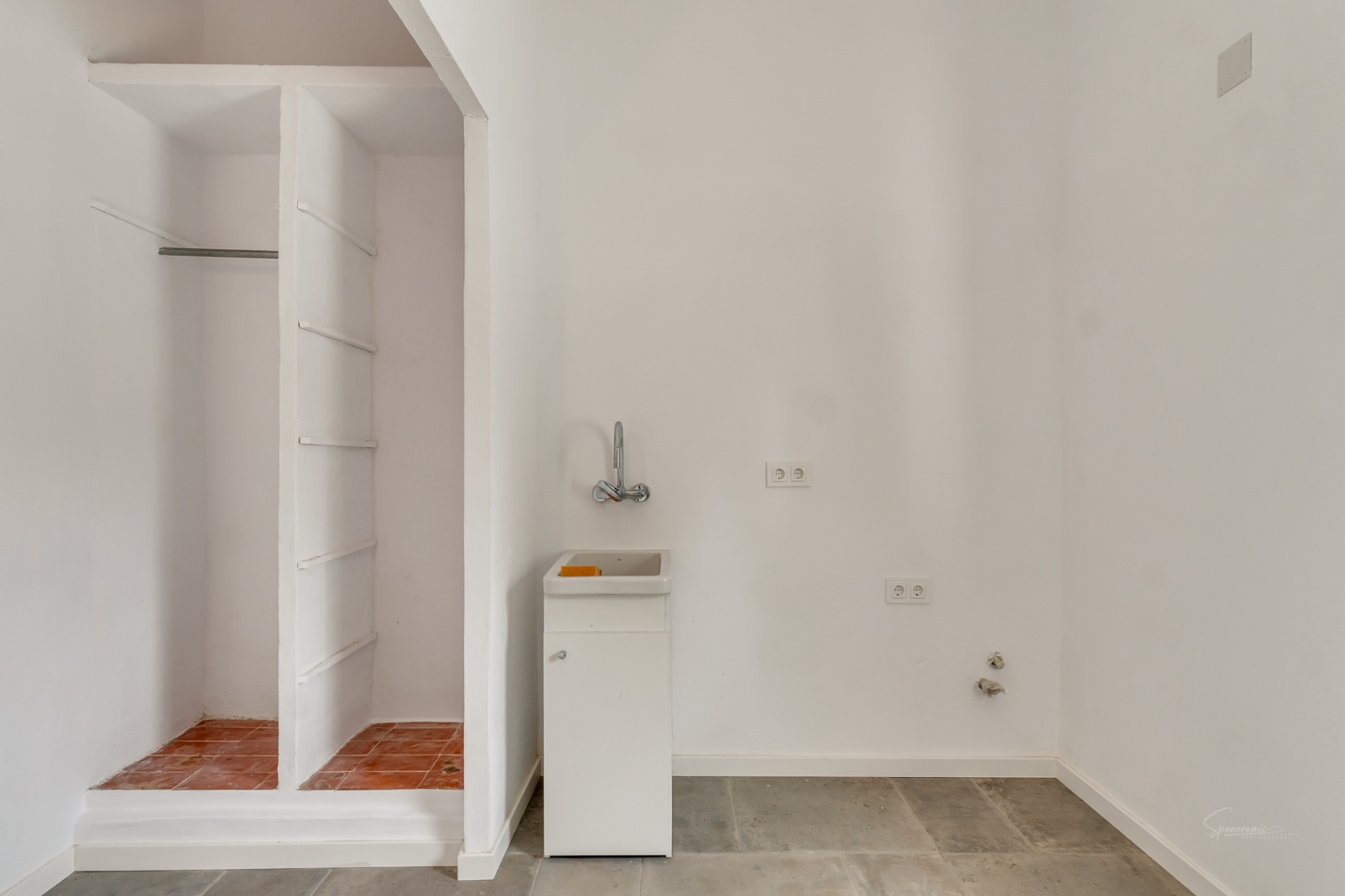 Utility area featuring built-in masonry shelving, a compact white sink with chrome fixtures, and durable grey stone flooring against white walls.