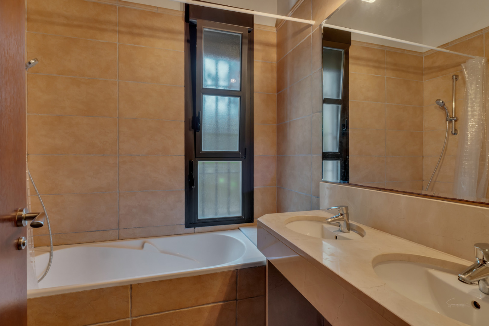 Bathroom featuring a double stone vanity, integrated bathtub, and earth-toned tiling complemented by a dark-framed window for natural light.