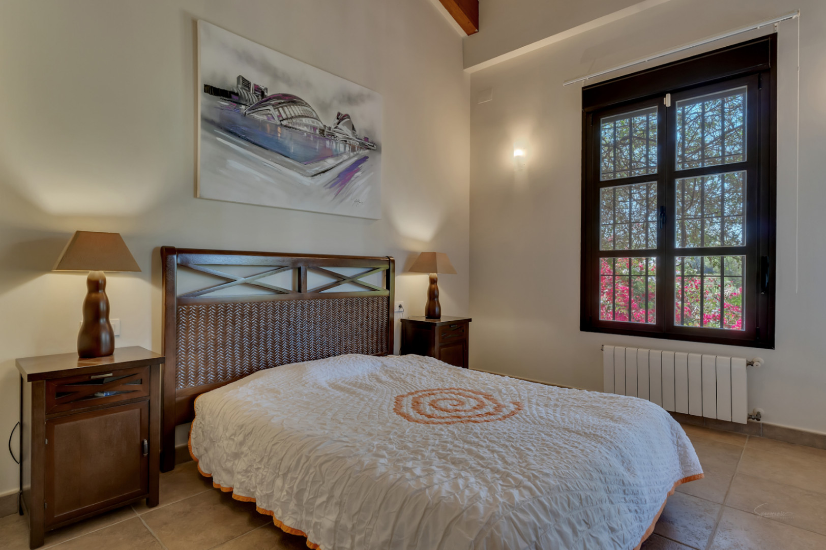 Bedroom featuring a dark wood bed with a woven headboard, exposed ceiling beams, and tile flooring. A large window offers views of the garden and bougainvillea.
