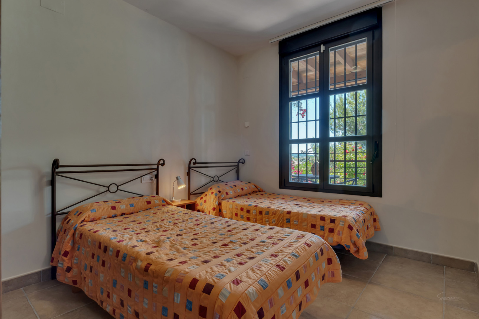 Bedroom featuring twin beds with wrought iron headboards and tiled flooring. A large window with security bars overlooks the lush garden greenery.