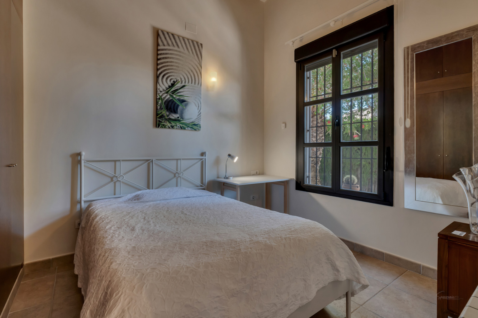 Bright bedroom featuring a white metal bed frame, a functional workspace, and a dark-framed window overlooking the garden. Finished with tiled floors.