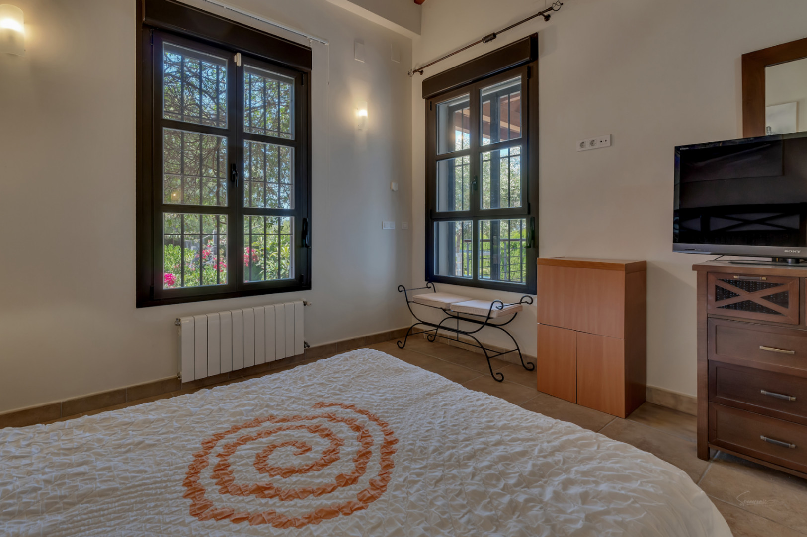 Bright bedroom featuring dark-framed windows with garden views, a wrought-iron bench, and classic wooden furniture on tiled flooring.