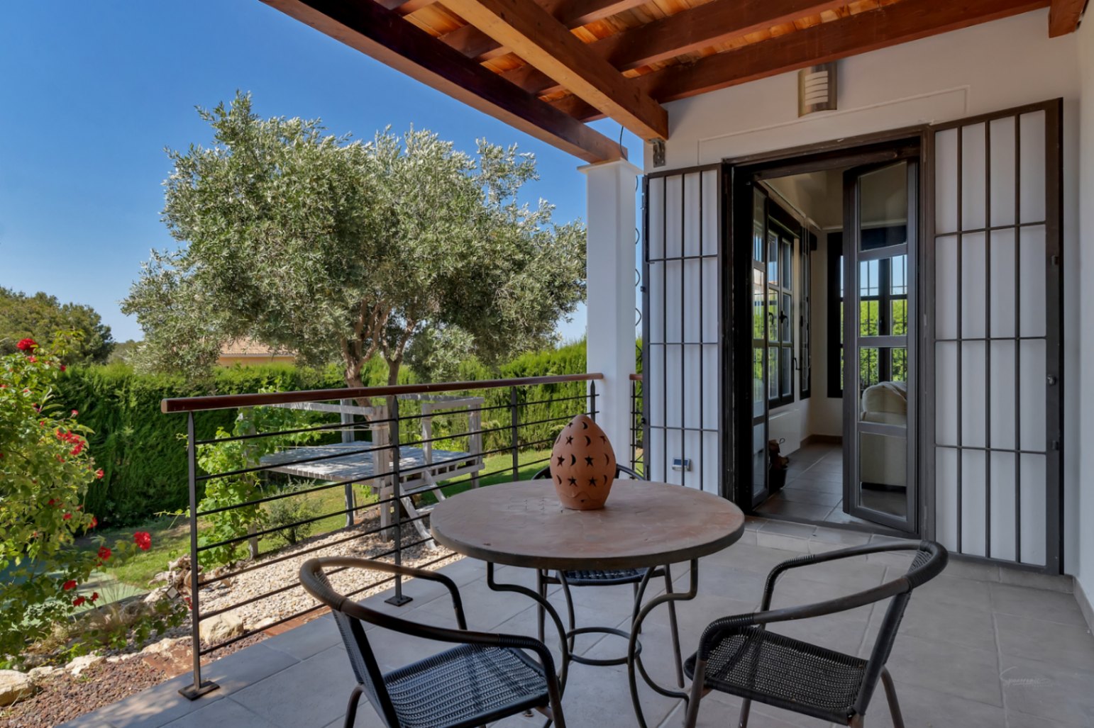 Covered terrace featuring exposed wooden beams, tiled flooring, and wrought iron furniture overlooking a Mediterranean garden with a mature olive tree.