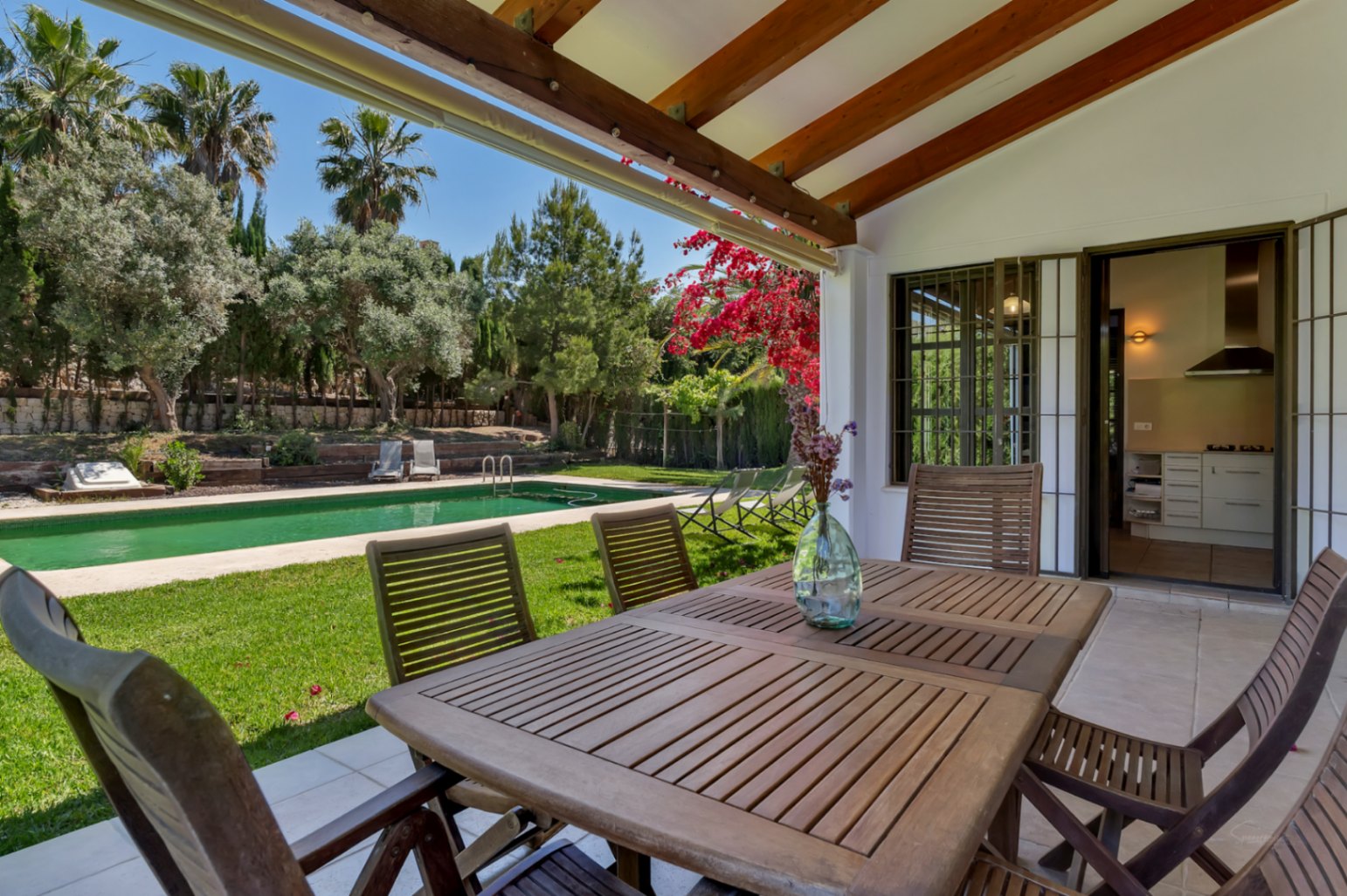 Covered terrace featuring wooden beams and an outdoor dining area, offering views of the Mediterranean garden and the rectangular swimming pool.
