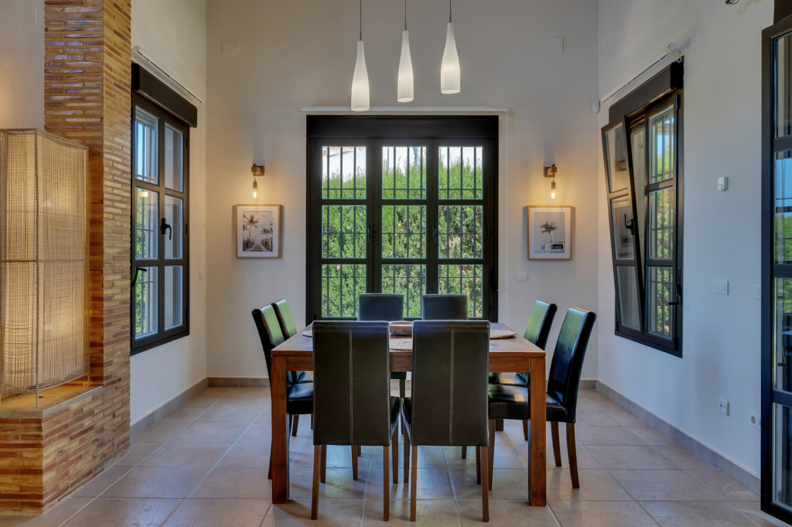 Dining area featuring a wooden table and black leather chairs under high ceilings. Dark-framed windows and a brick pillar accent the bright, tiled space.