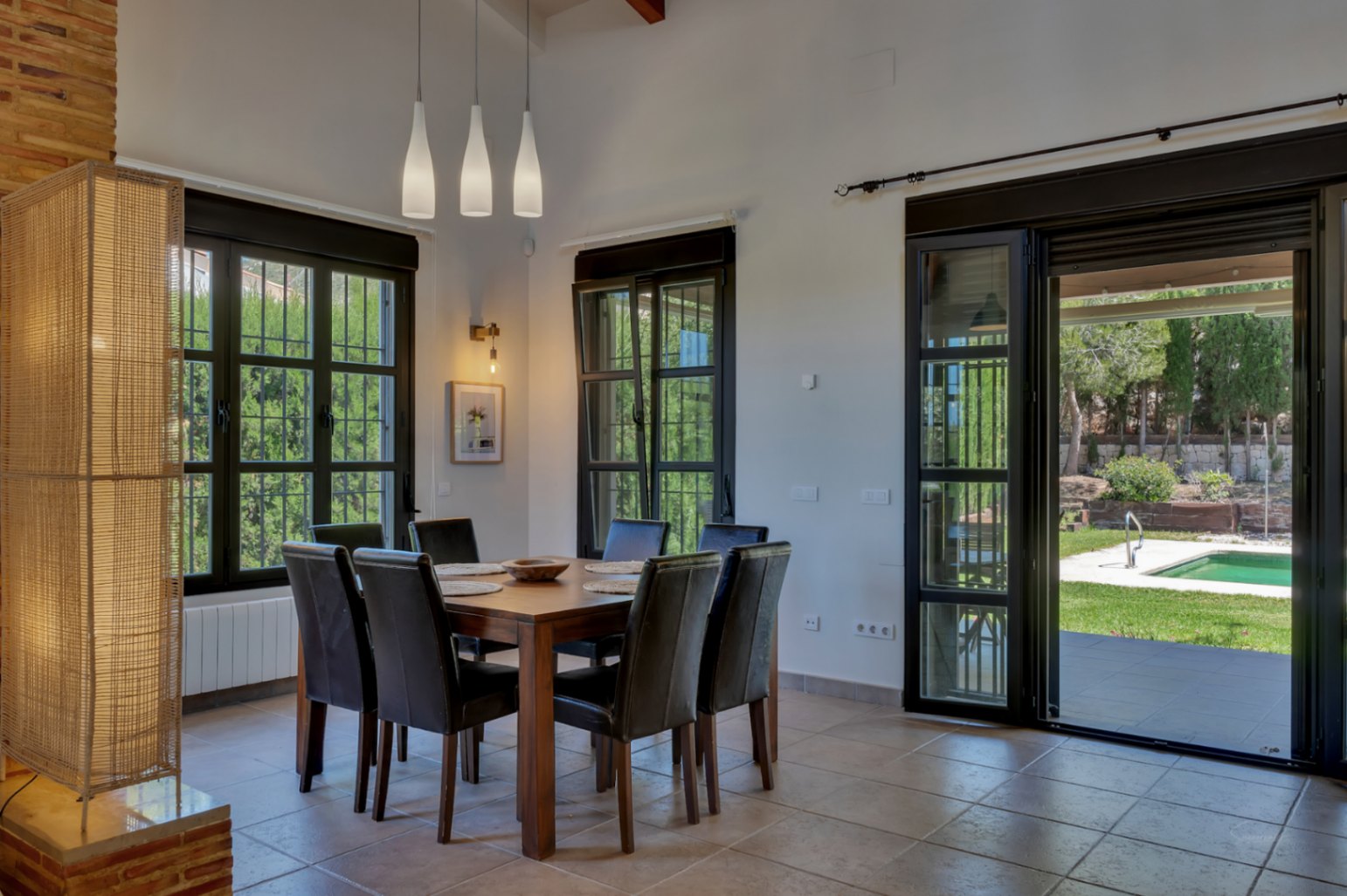 Dining area featuring a wooden table and dark leather chairs, with direct access to the garden and pool through large black-framed glass doors.