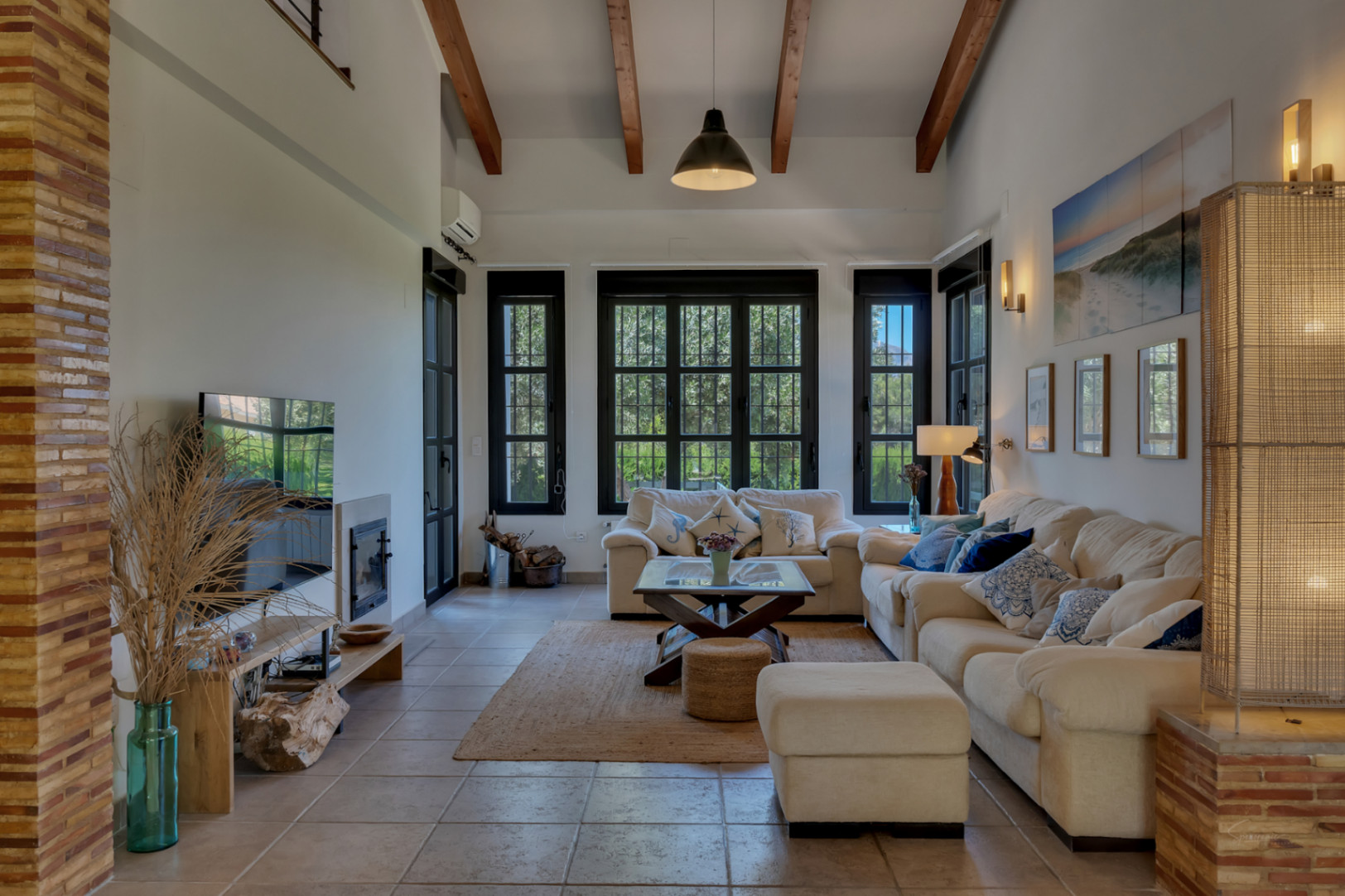 Elegant living room featuring high ceilings with exposed wooden beams, an integrated fireplace, and large black-framed windows that offer views of the garden.