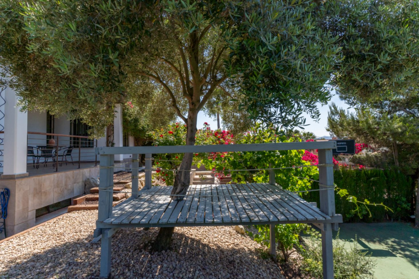 Elevated grey wooden platform built around a mature tree, set within a landscaped Mediterranean garden with gravel and flowering shrubs.