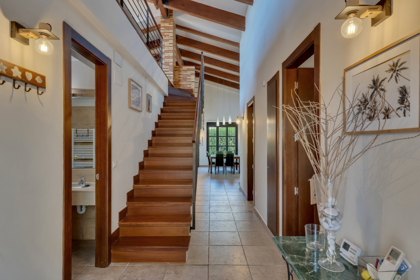 Entrance hallway featuring high ceilings with exposed wooden beams, a solid wood staircase, and tiled flooring leading towards the bright dining area.