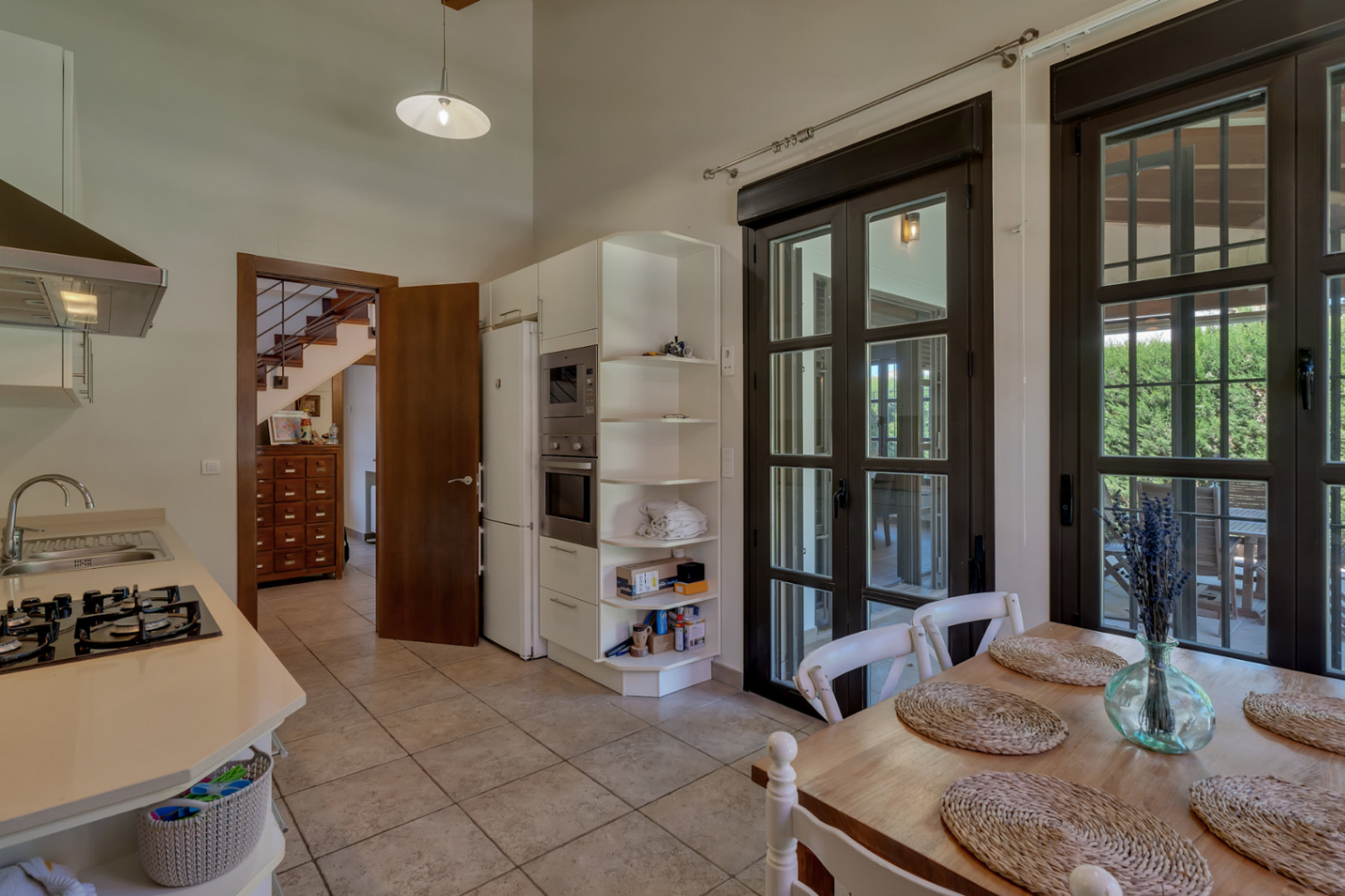 Equipped kitchen featuring white cabinetry, integrated appliances, and a dining area. It offers high ceilings, tiled floors, and direct terrace access via large glass doors.