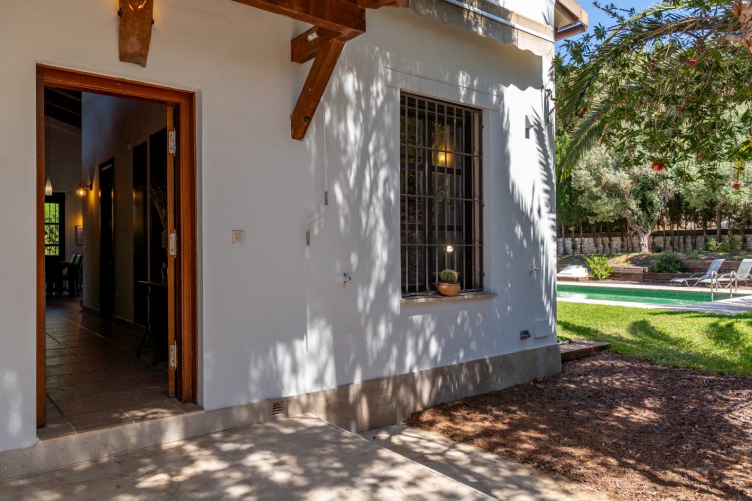 Exterior entrance featuring an open wooden door, white facade with timber beams, and a view towards the garden, swimming pool, and lawn.