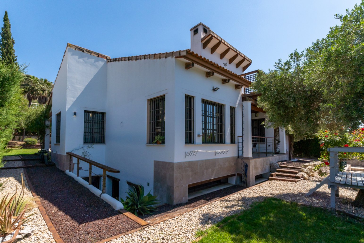 Exterior view of the villa featuring white walls, wooden eaves, and stone cladding. The landscaped garden includes gravel paths and access to the lower level.