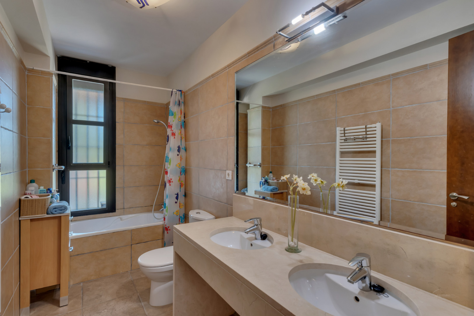 Full bathroom featuring a double vanity with stone countertop, large mirror, integrated bathtub, and neutral sand-toned ceramic tiling throughout.