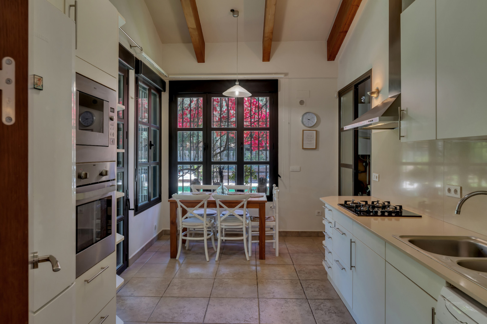Functional kitchen with high ceilings, exposed wooden beams, integrated appliances, and a dining area with views of the garden through dark-framed windows.