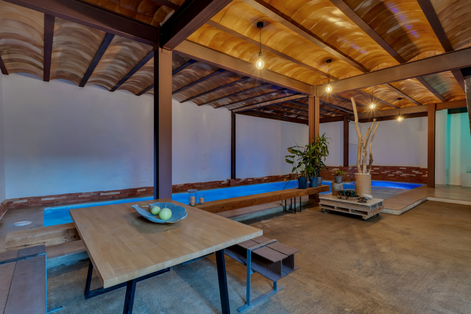 Indoor heated pool featuring blue ambient lighting and a vaulted terracotta tile ceiling with industrial beams. The area includes a wooden dining set and concrete flooring.