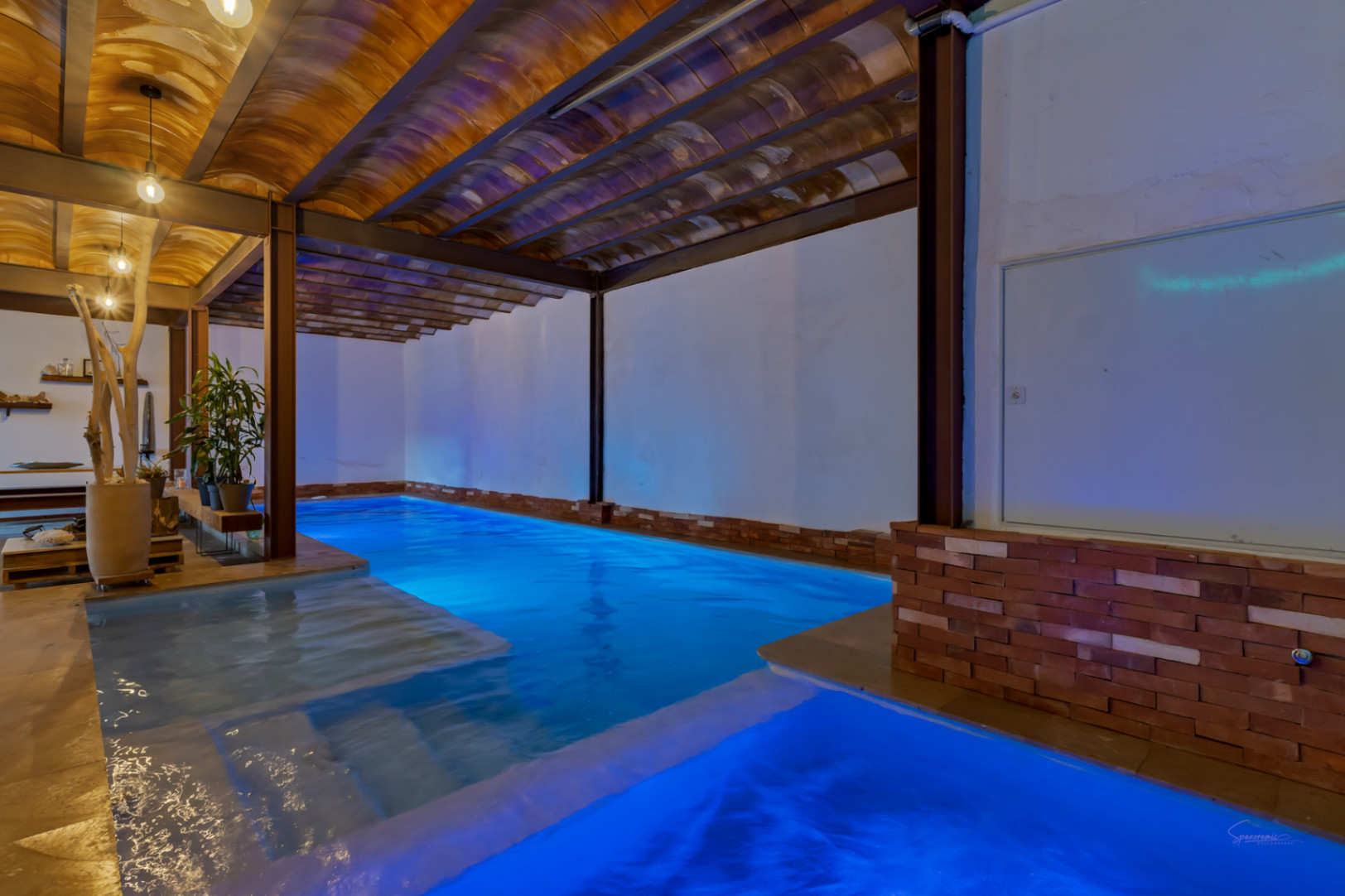 Indoor heated pool featuring blue LED lighting, vaulted terracotta ceilings, and exposed steel beams for a blend of rustic and industrial design.