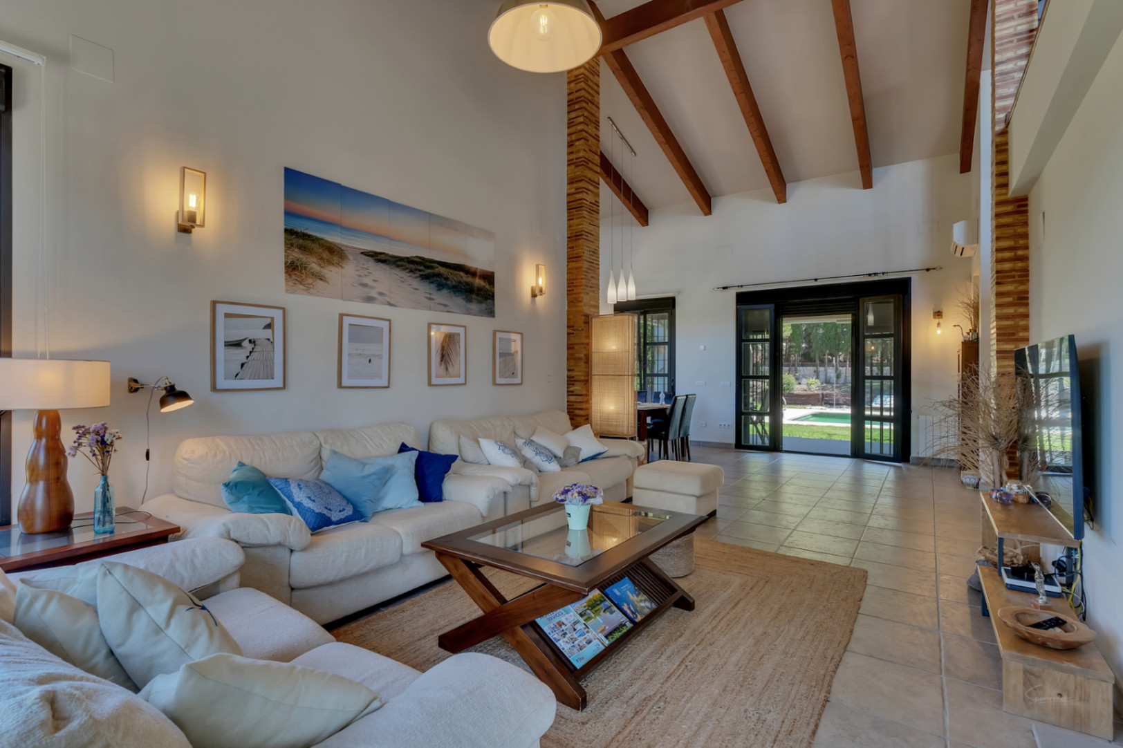 Living area with double-height ceilings, exposed wooden beams, and large glass doors opening directly onto the terrace and garden pool.