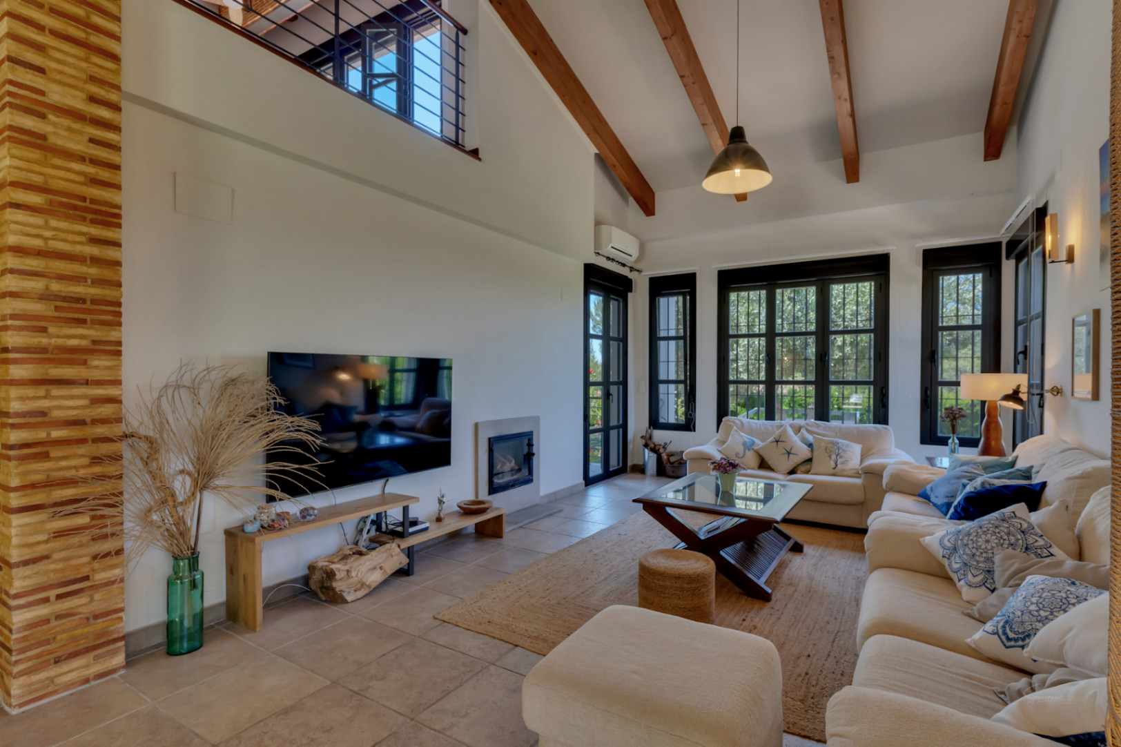 Living room featuring high vaulted ceilings with wooden beams, an integrated fireplace, and large black-framed windows offering garden views.