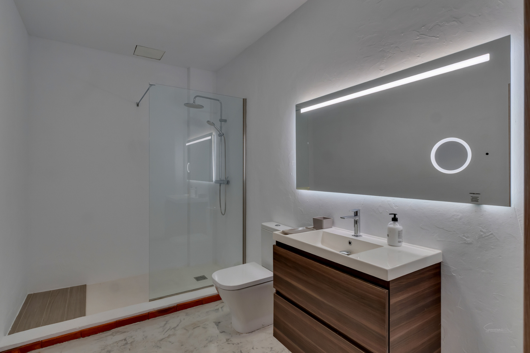 Modern bathroom featuring a floating wood-finish vanity, integrated sink, and large backlit mirror. Includes a walk-in shower with glass partition and textured white walls.