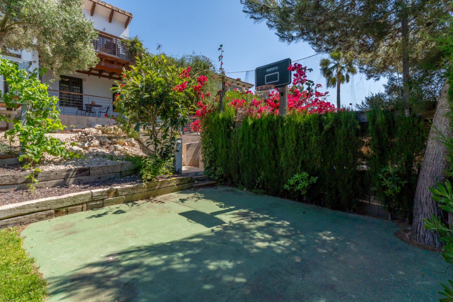 Private outdoor basketball court featuring a green hardcourt surface, hoop, and wooden sleeper retaining walls, bordered by mature trees and bougainvillea.