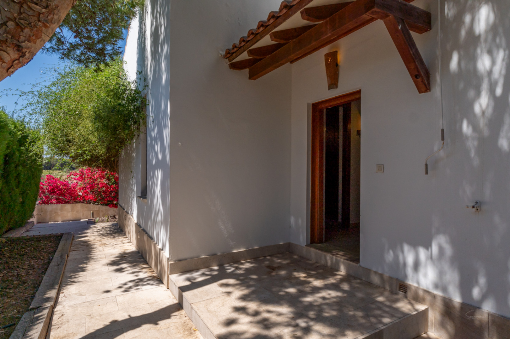Side entrance featuring a wooden beam porch with clay tiles, natural stone flooring, and a garden path lined with Mediterranean flora.