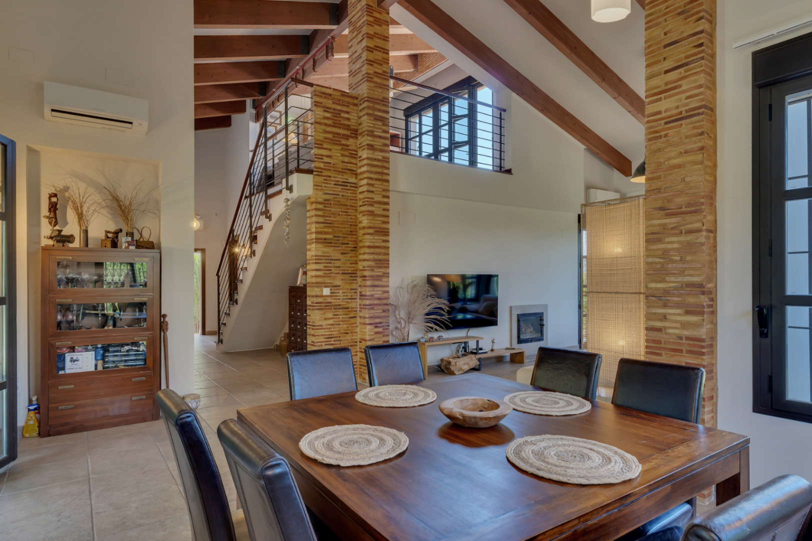 Spacious living and dining area with high ceilings, exposed wooden beams, brick pillars, and a staircase leading to the open upper-level office space.