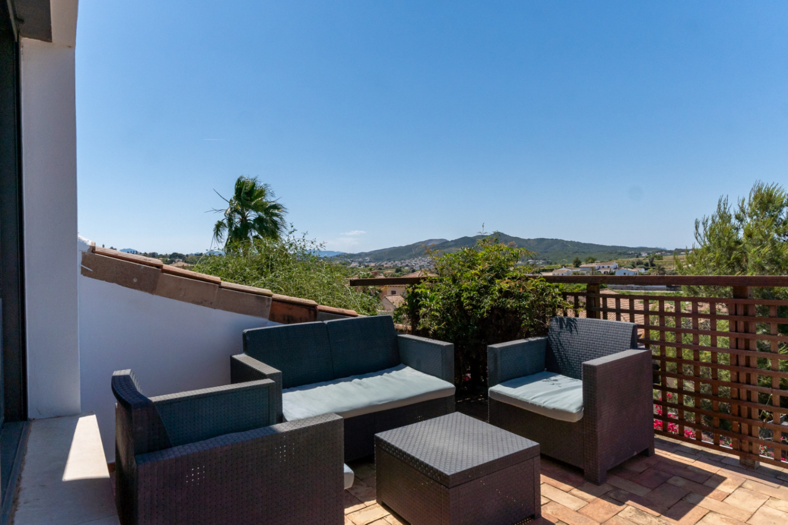 Upper terrace featuring a synthetic rattan lounge set and terracotta tiling, overlooking the valley and distant mountains under a clear sky.
