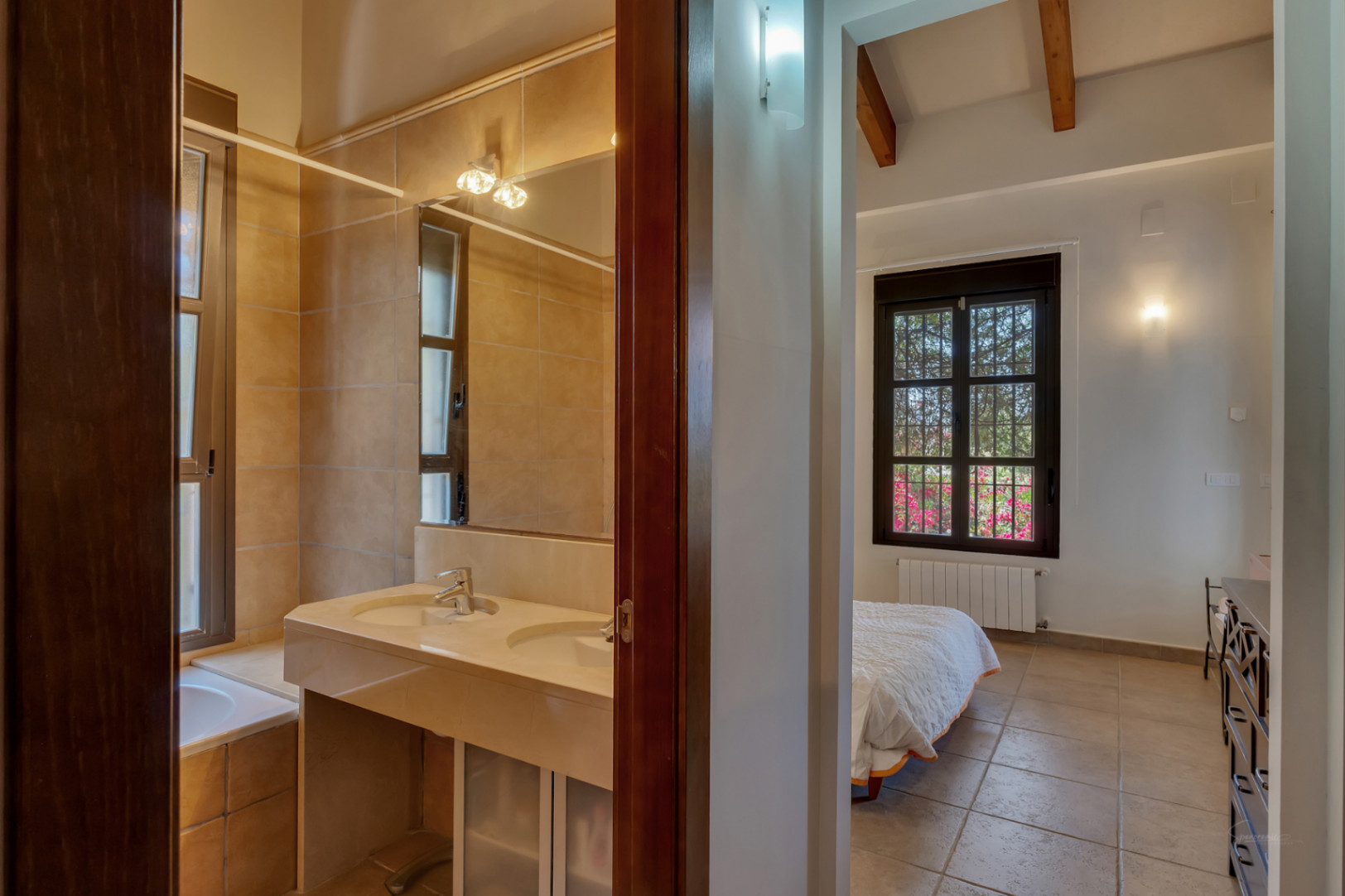 View from the hallway showing an en-suite bathroom with a double stone vanity and a bedroom featuring exposed wooden beams and garden views.