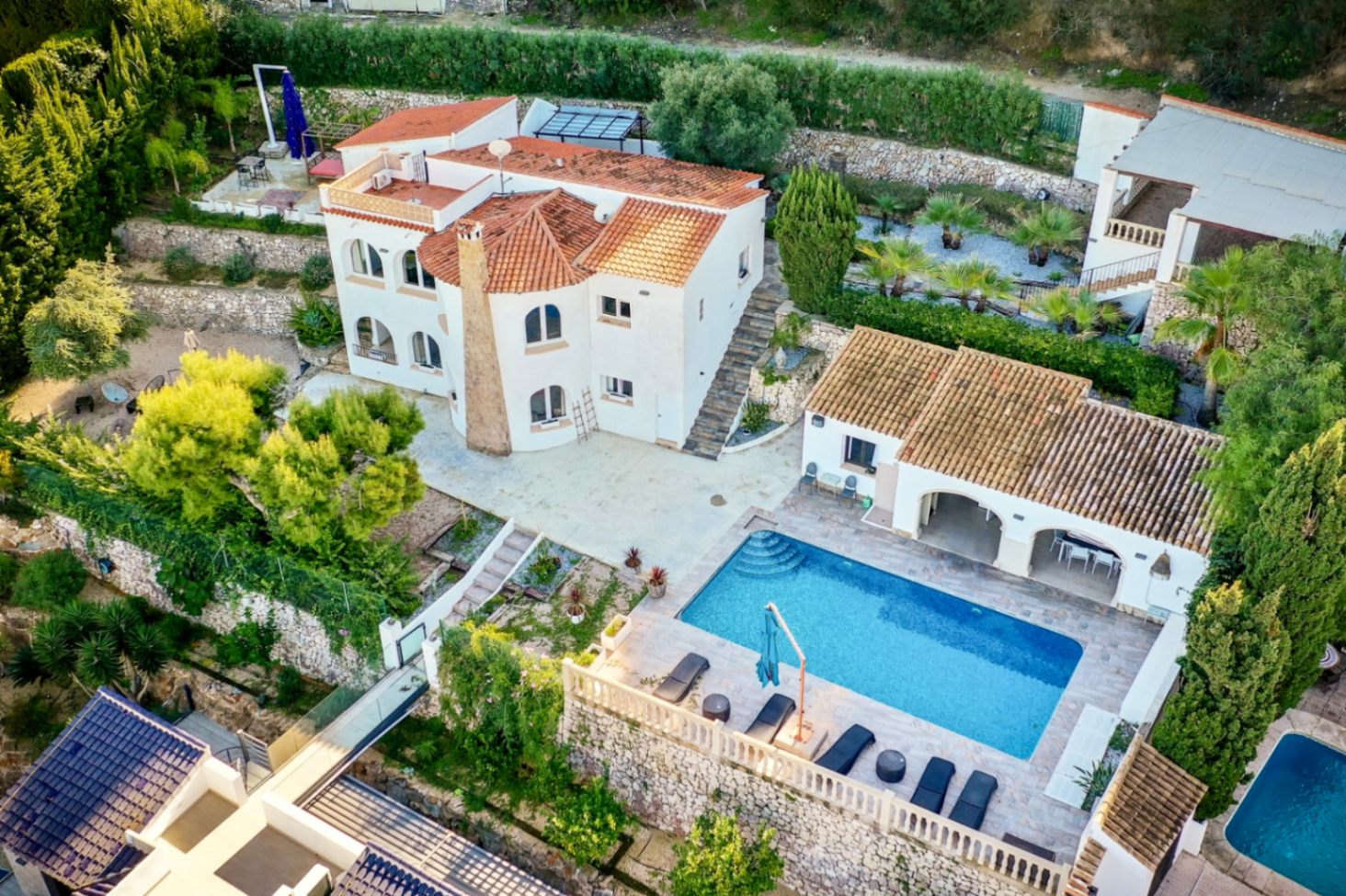 Aerial view of the Mediterranean villa featuring a rectangular pool, stone-paved sun terrace, outdoor kitchen, and terraced gardens with native vegetation.
