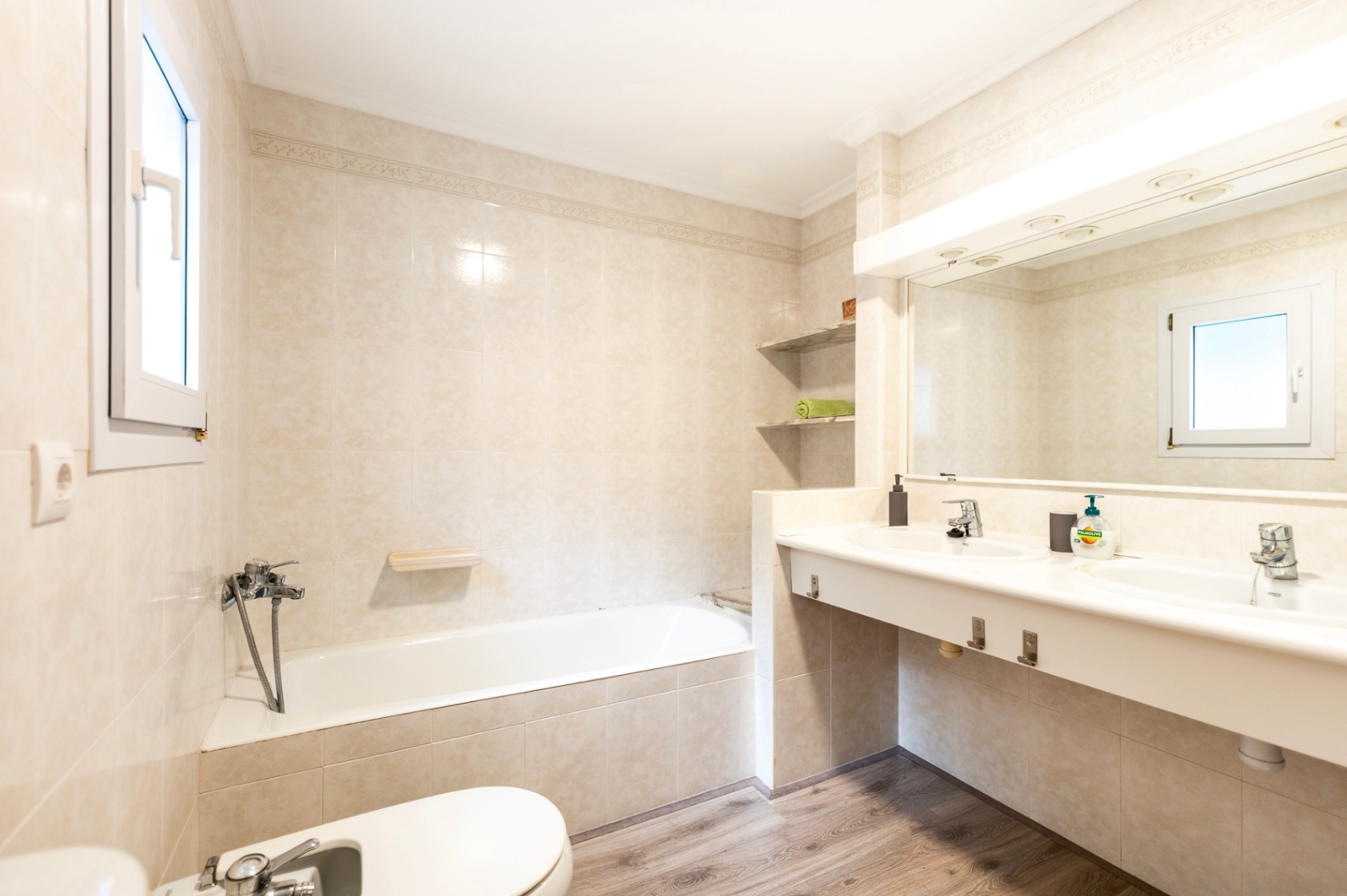 Bathroom featuring a built-in bathtub, double vanity, and wood-effect flooring. The space is finished with cream tiles, a large mirror with integrated lighting, and natural light.