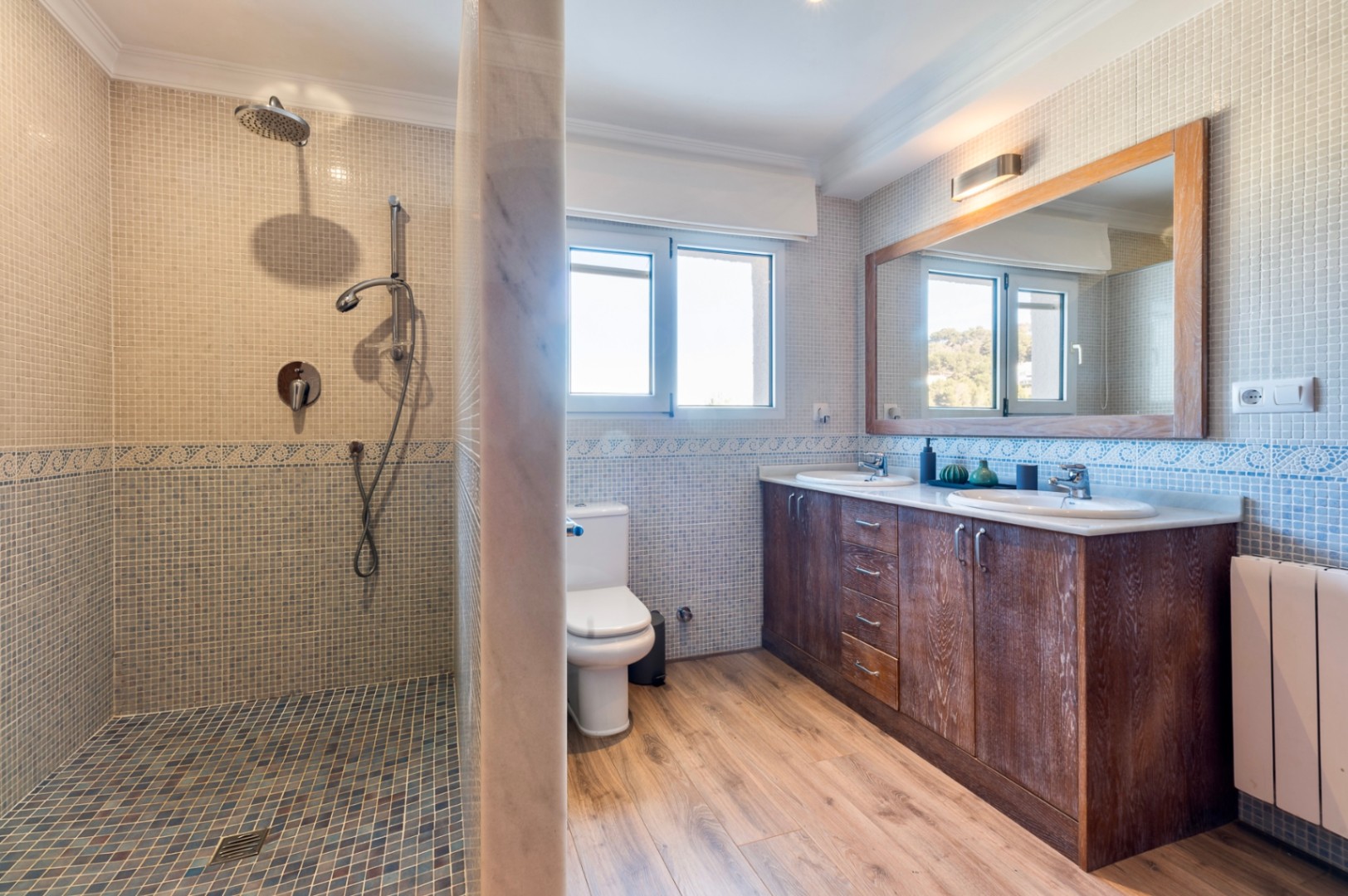 Bathroom featuring a mosaic-tiled walk-in shower, double vanity with dark wood cabinetry, and a large wooden-framed mirror under natural light.
