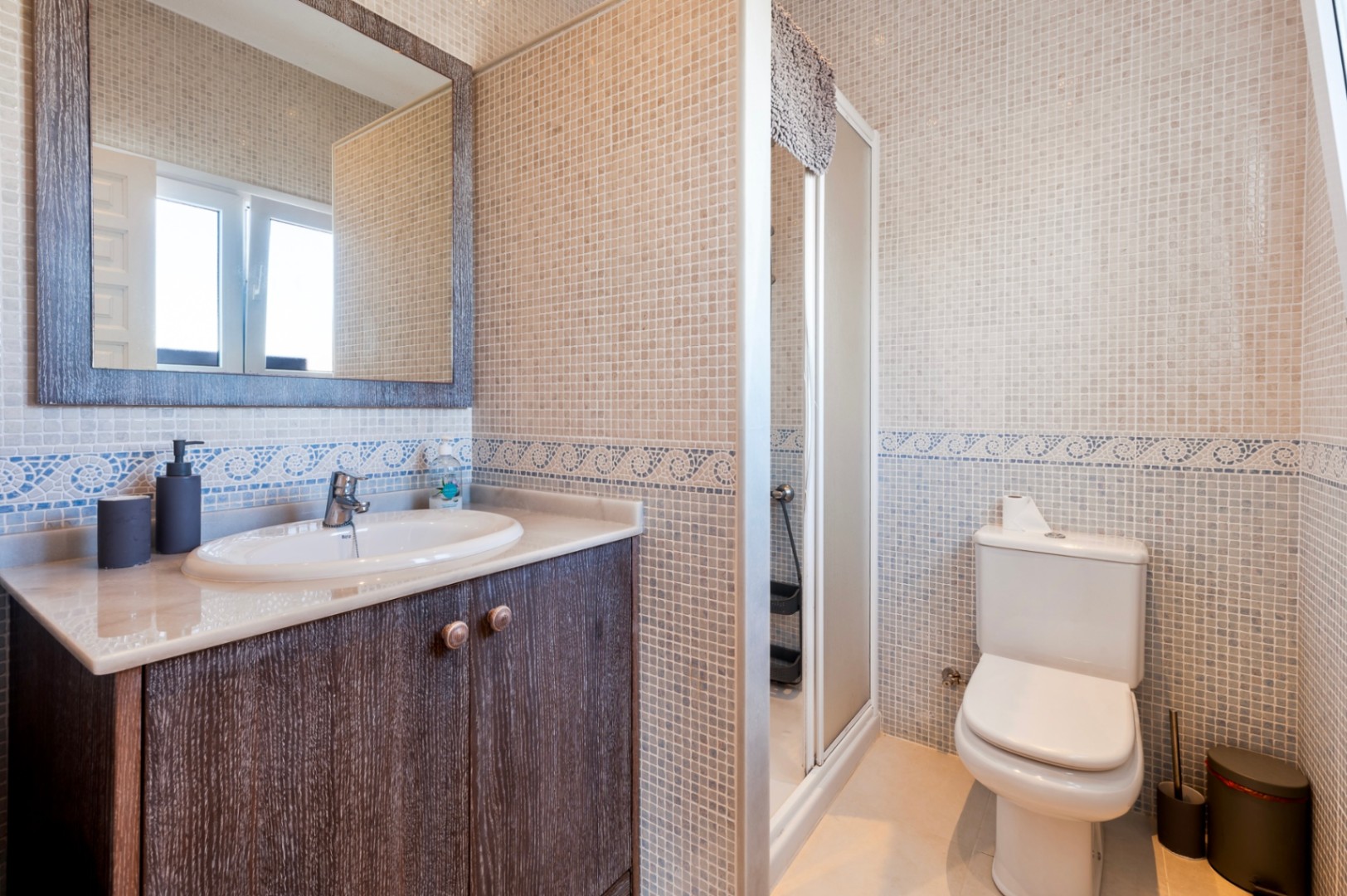 Bathroom featuring cream mosaic tiles with a blue wave border, a dark wood vanity with marble top, and a glass-enclosed shower.
